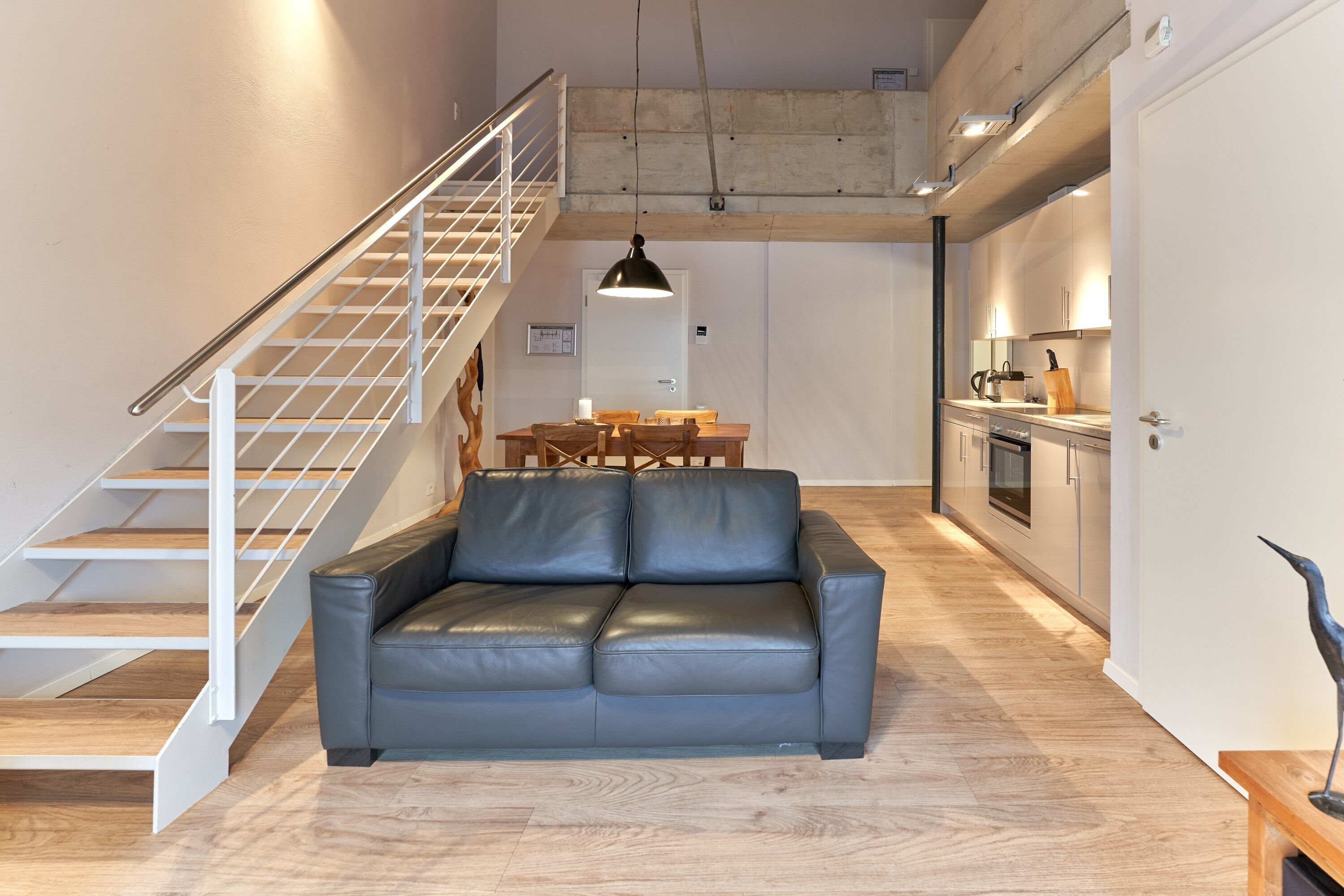 A modern living room with a leather sofa, wooden flooring, a staircase, and a kitchen area.