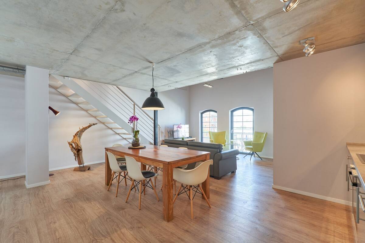 A spacious loft with a high ceiling, a dining area with a wooden table and chairs, a staircase, and a living area with a sofa and chairs.