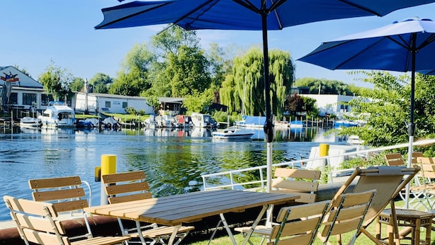 A lakeside patio with wooden tables and chairs under blue umbrellas, surrounded by boats and greenery. - havelblau FERIENLOFTS (Brandenburg an der Havel)