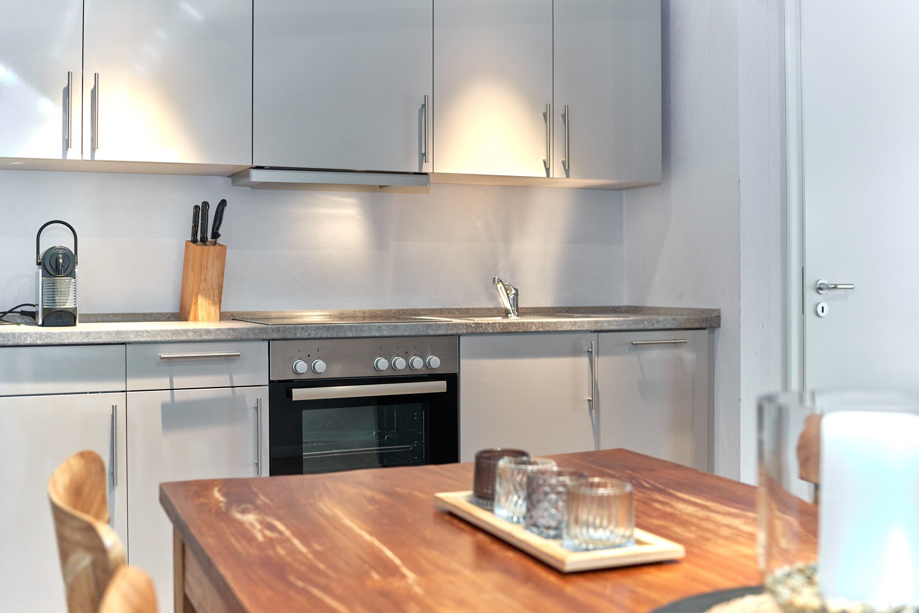 Una cocina moderna con armarios blancos, horno empotrado y una mesa de comedor de madera con vasos y una bandeja.