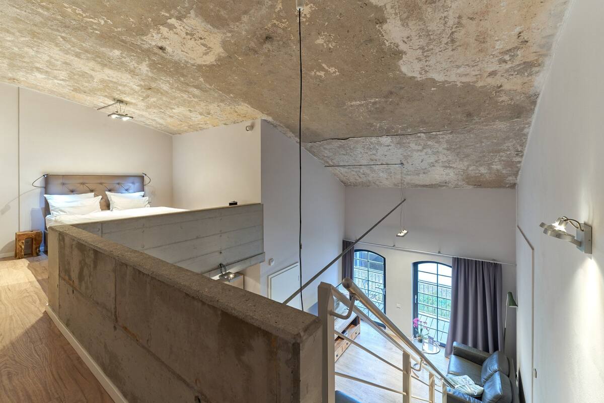 A high-ceilinged room with a concrete ceiling, a bed, a staircase, and a view of the outdoors through a window.