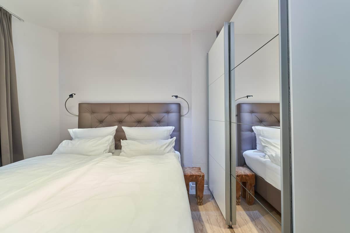 A modern bedroom with a large bed, a wooden stool, and a mirror on the wall.