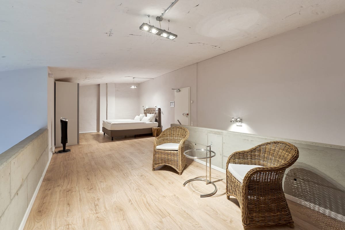 A hotel room with a wooden floor, a bed, two wicker chairs, a small table, and a glass partition.