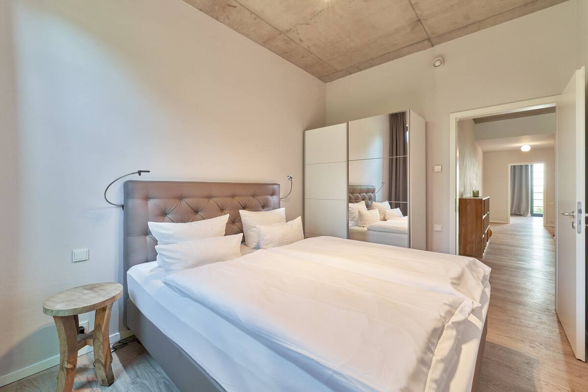 A modern bedroom with a large bed, a wooden stool, a mirror, and a wardrobe.