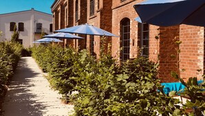 A pathway lined with lush greenery and blue umbrellas, leading to a brick building with arched windows. - havelblau FERIENLOFTS (Brandenburg an der Havel)