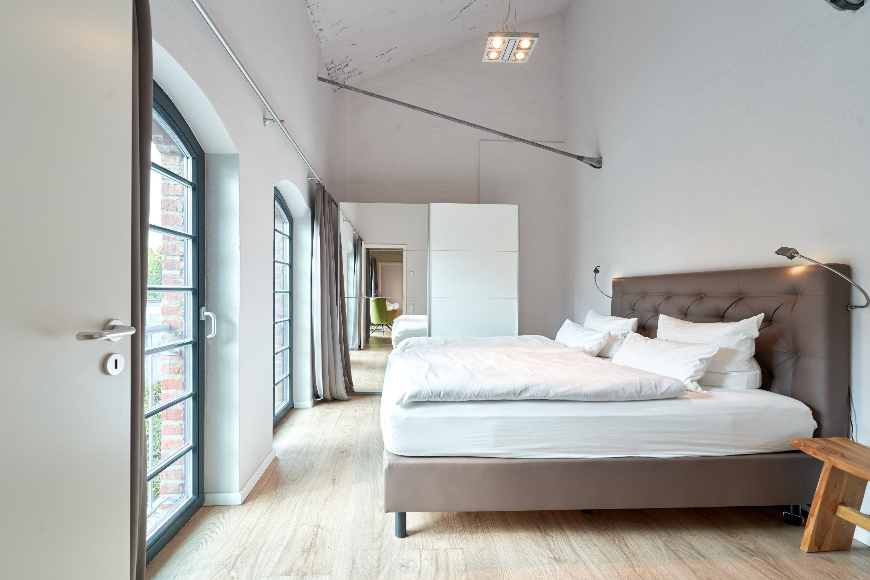 A modern bedroom with a large bed, a wooden bench, and a view of a cityscape through large windows.