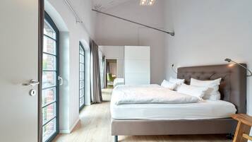 A modern bedroom with a large bed, a wooden bench, and a view of a cityscape through large windows.