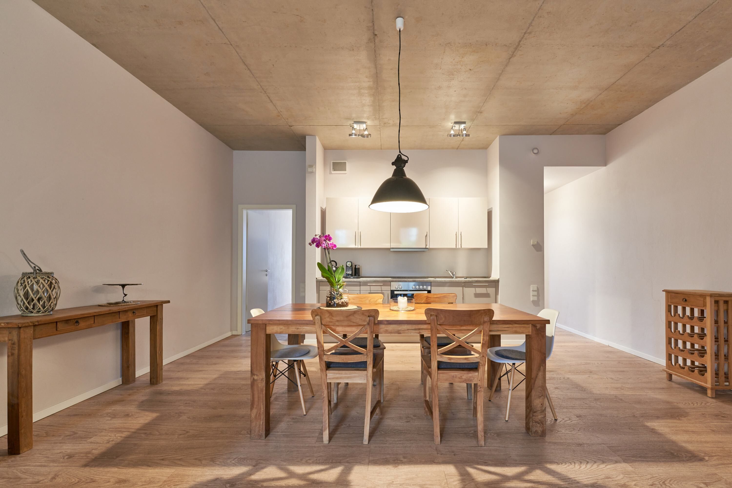 Un comedor espacioso con una mesa y sillas de madera, una lámpara colgante y una cocina al fondo.