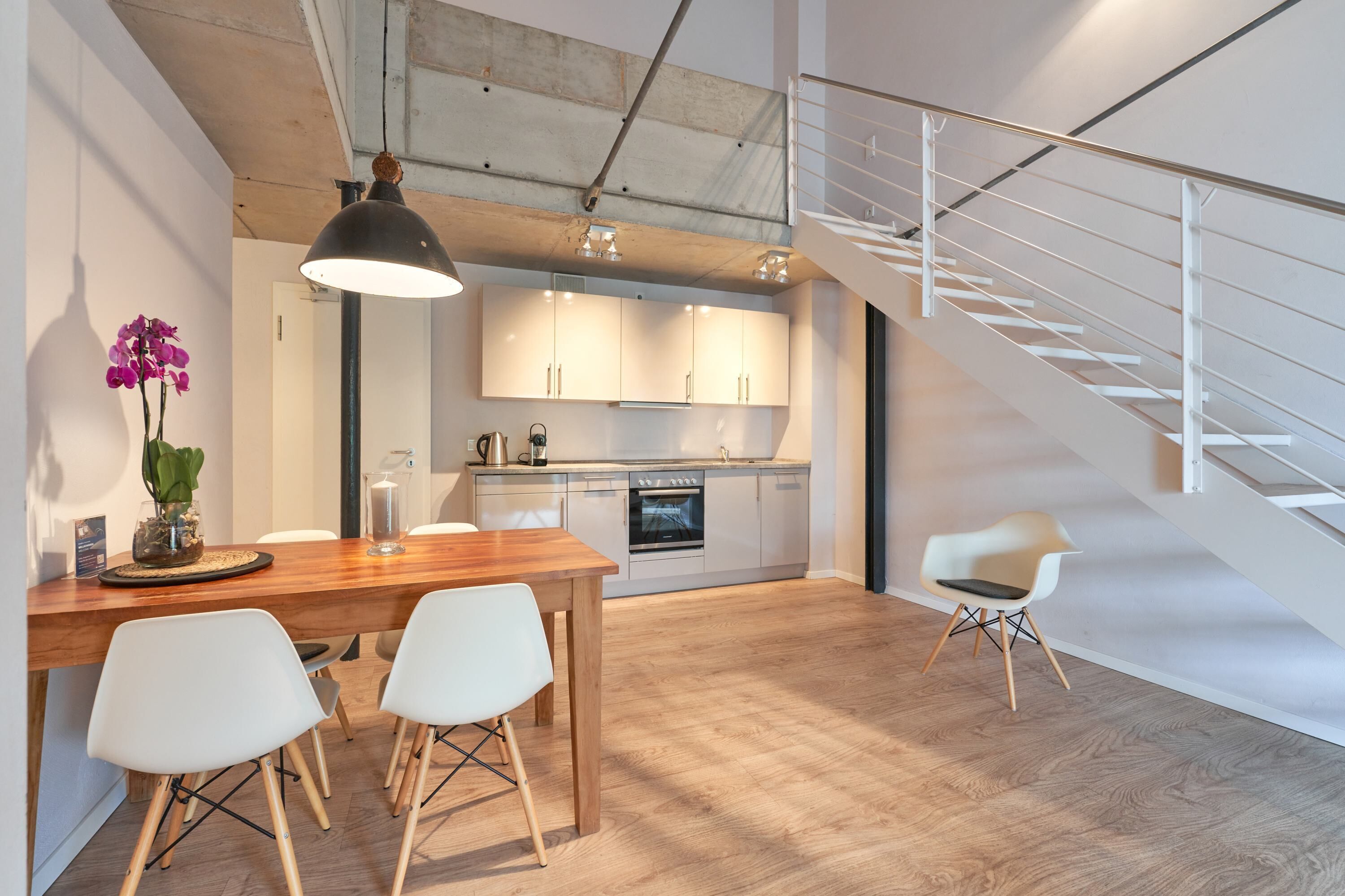 Una cocina moderna con armarios blancos, una mesa de comedor de madera y dos sillas blancas. Hay una escalera a la derecha y una lámpara colgante sobre la mesa.