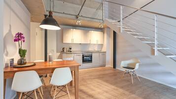 Una cocina moderna con armarios blancos, una mesa de comedor de madera y dos sillas blancas. Hay una escalera a la derecha y una lámpara colgante sobre la mesa.