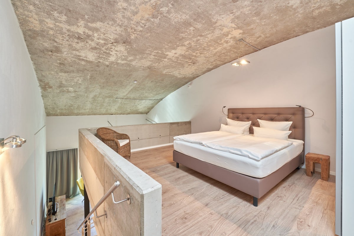 A bedroom with a large bed, a wooden floor, a concrete ceiling, and a view of the sky.