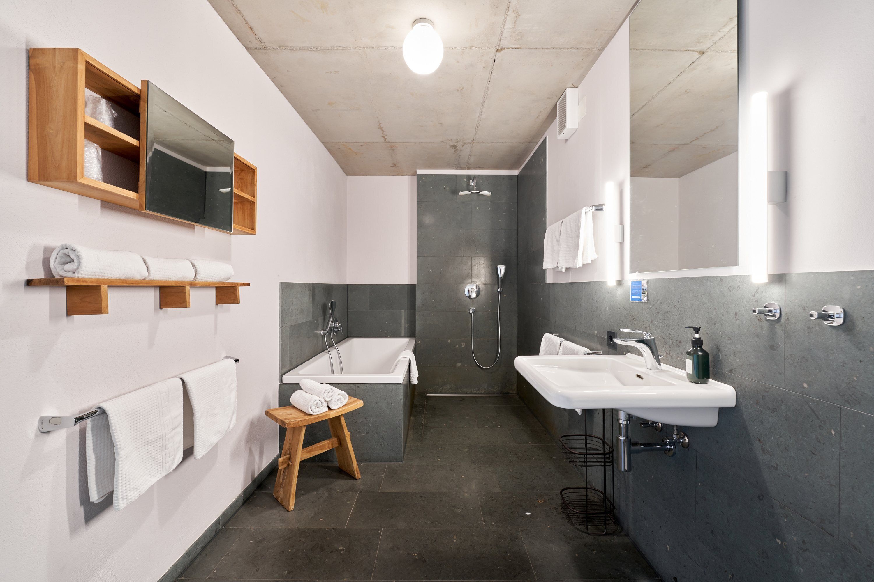Ein modernes Badezimmer mit einer Badewanne, zwei Waschbecken, einer Dusche, Holzregalen und einem kleinen Hocker.