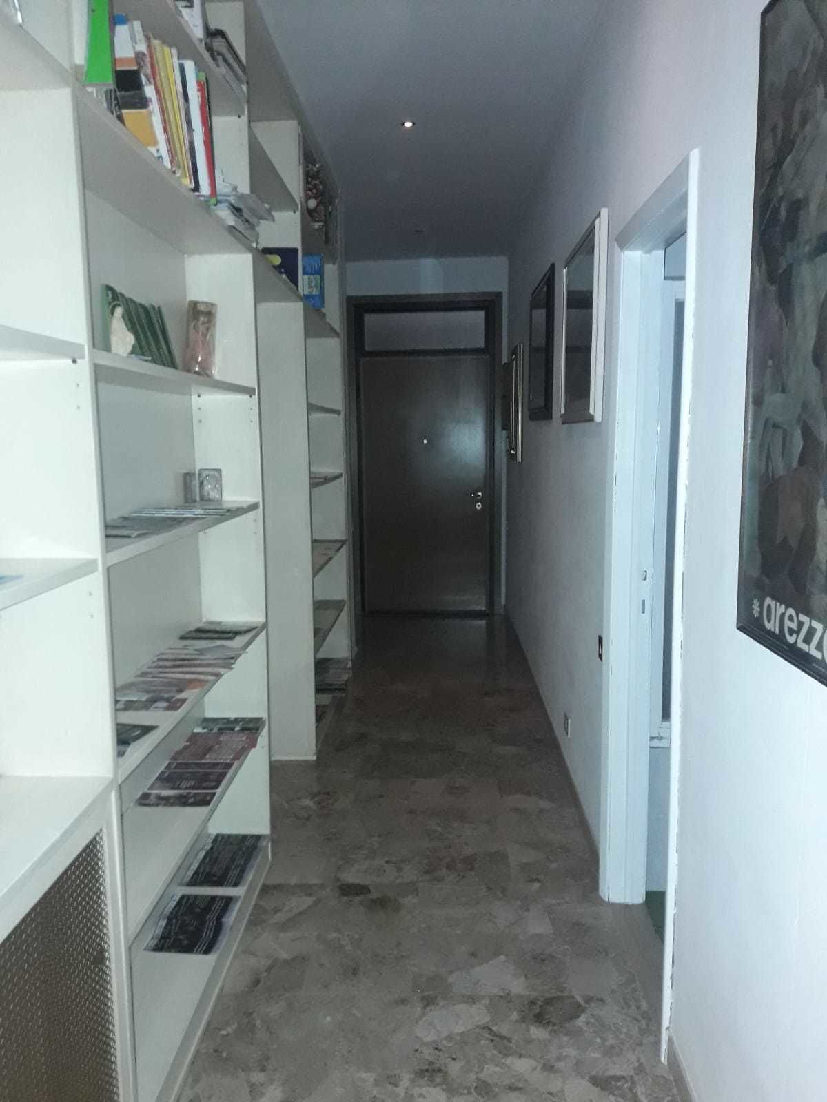 A hallway with a bookshelf filled with books and magazines, a door at the end, and a poster on the wall.