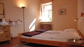 A bedroom with a wooden bed, a bedside table with a book, a radiator, a window with curtains, a floor lamp, and a dresser with towels.