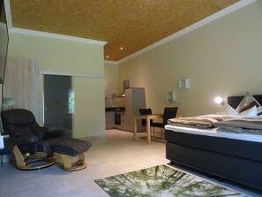 Habitación de hotel moderna con techo de madera, sofá cama, mesa de comedor con sillas, zona de cocina y vistas al exterior.