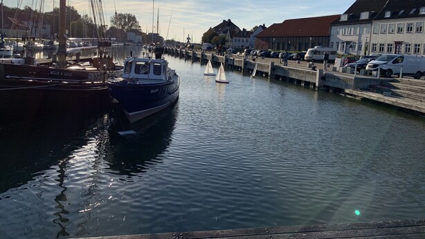 Un port de plaisance avec des bateaux amarrés, un voilier sur l’eau et des bâtiments le long de la rive.