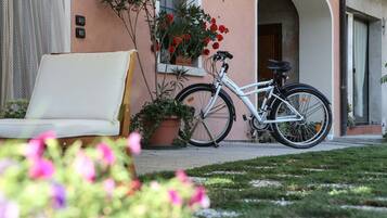 Una bicicleta blanca estacionada frente a un edificio de fachada rosa, una silla blanca al aire libre y macetas con flores rojas.
