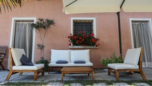 Un patio con un sofá blanco, dos sillas de madera y una mesa de madera. Hay plantas en macetas y una cesta de flores colgante en la pared.