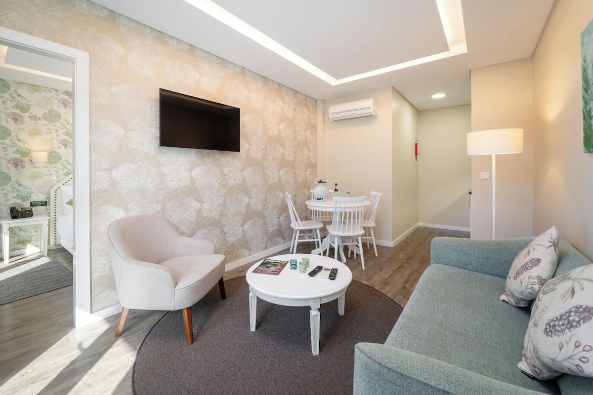 A modern living room with a white sofa, a round coffee table, a dining area with a white table and chairs, a mounted TV, and an air conditioning unit.
