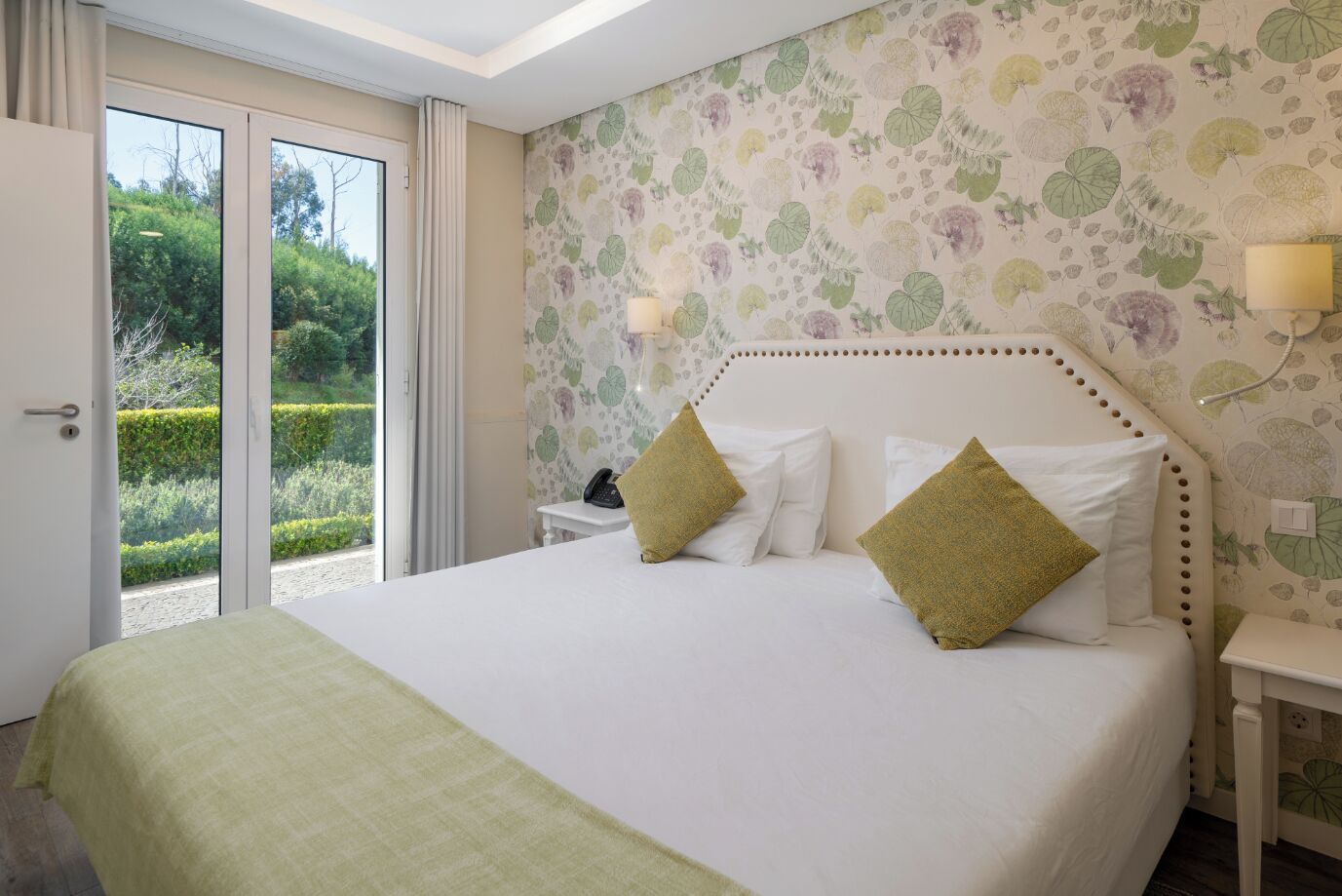 Une chambre à coucher avec un grand lit, deux tables de chevet, une commode et une vue sur un jardin, accessible par une porte coulissante.
