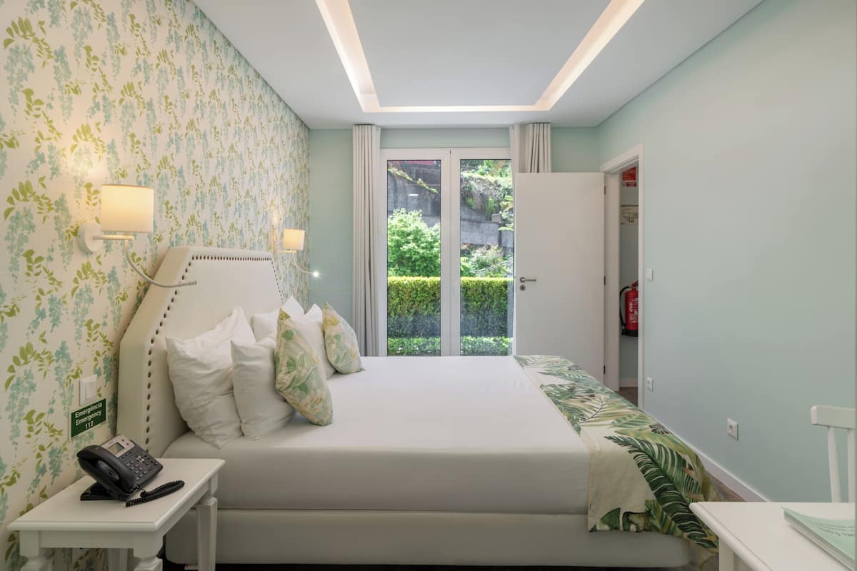 A bedroom with a bed, bedside table, lamp, and a view of greenery through a door.