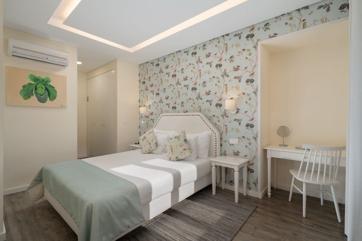 A bedroom with a bed, bedside table, chair, and a desk with a chair. There is a wall-mounted air conditioner and a painting on the wall.