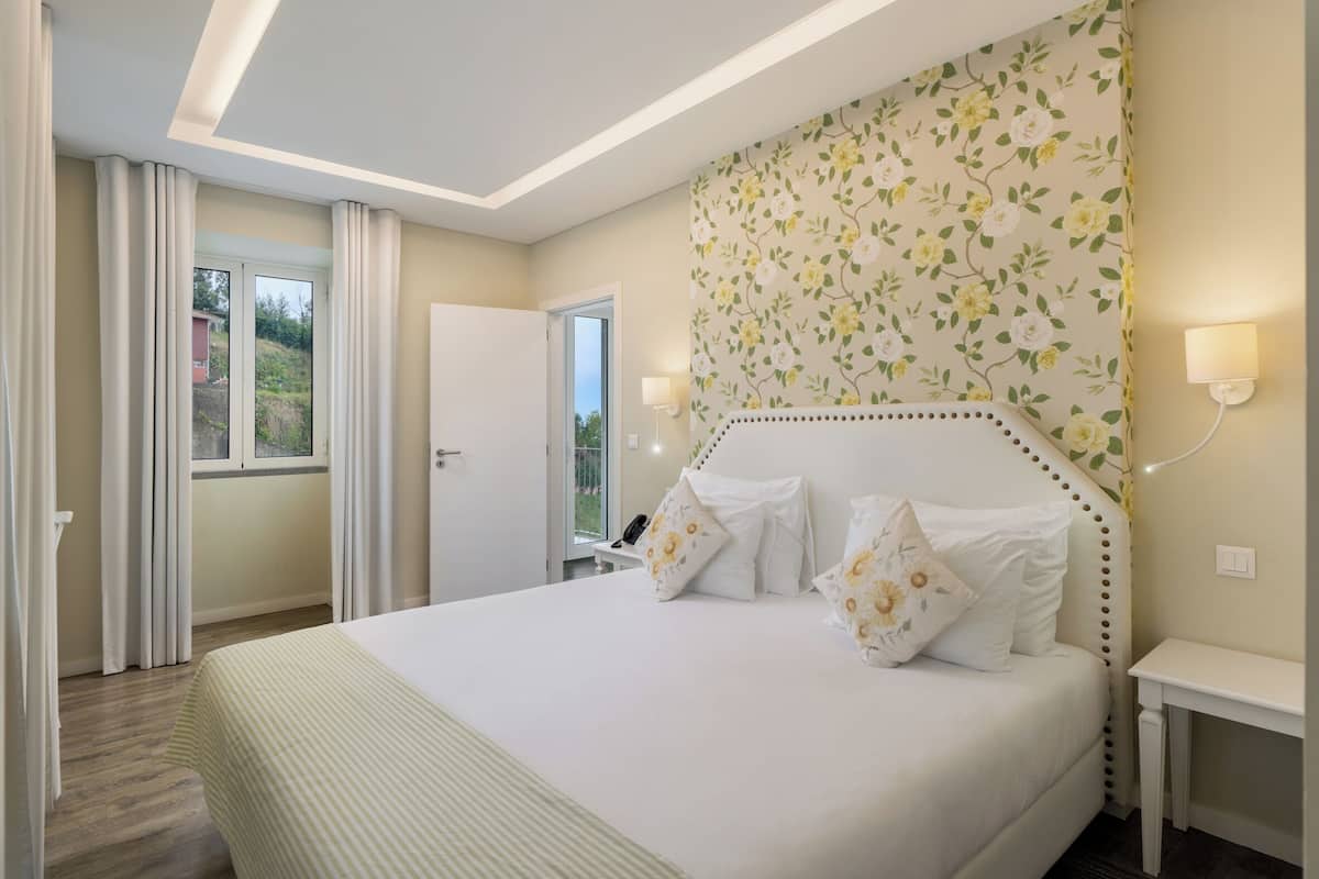 A hotel room with a large bed, floral wallpaper, a bedside table with a lamp, and a view of the outdoors through a window.