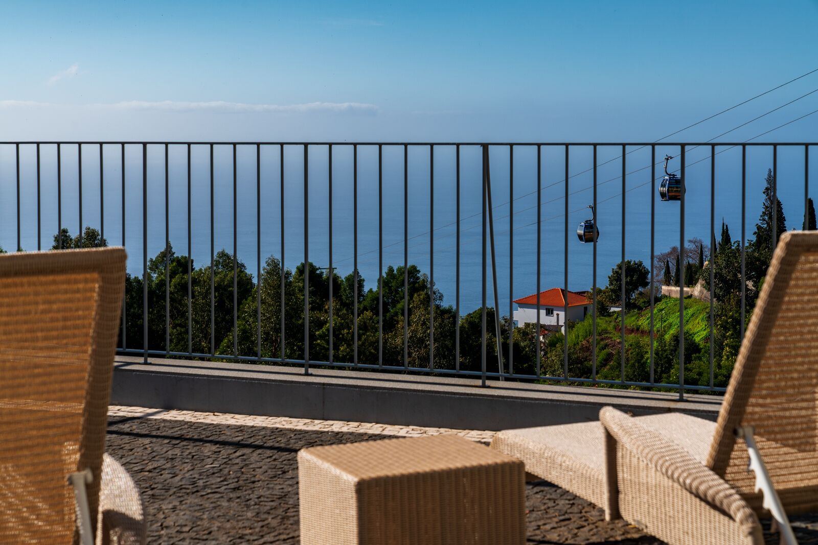 Un balcon avec des fauteuils en osier, une rambarde en métal et une vue sur la mer et un téléphérique.