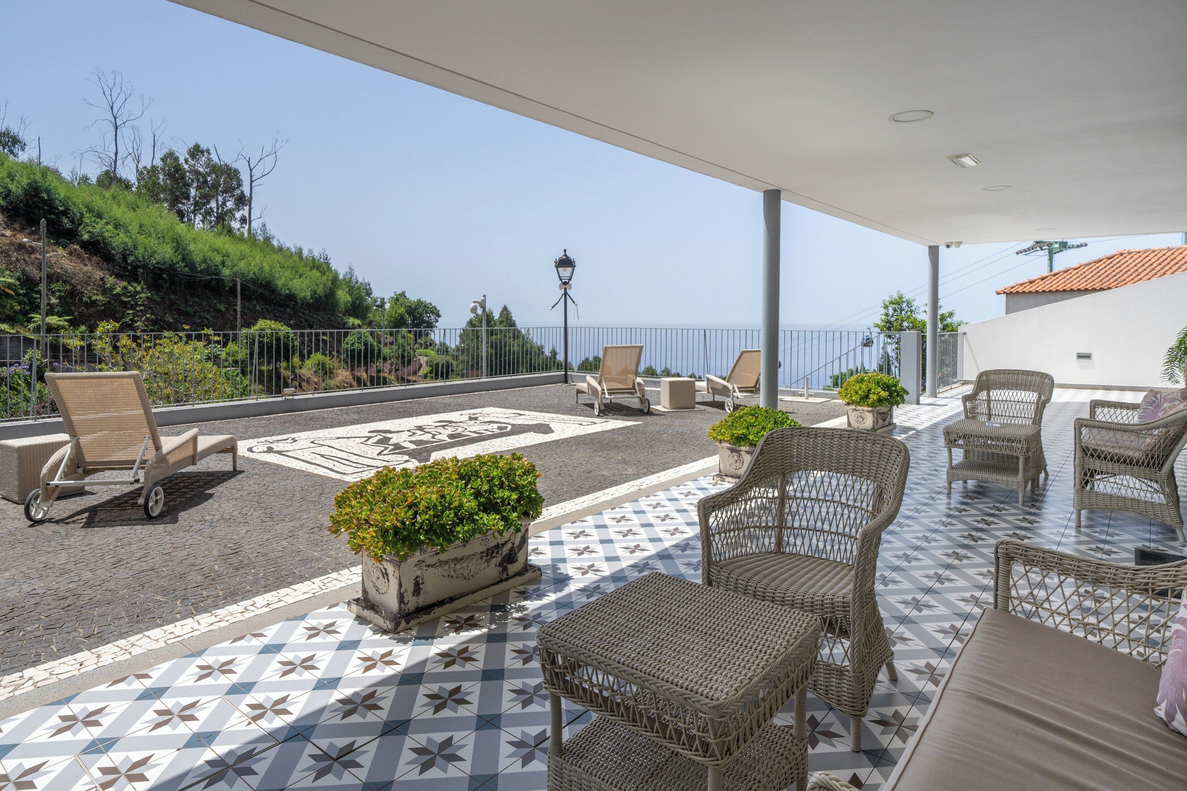 A covered outdoor seating area with wicker furniture and potted plants, overlooking a scenic hillside.