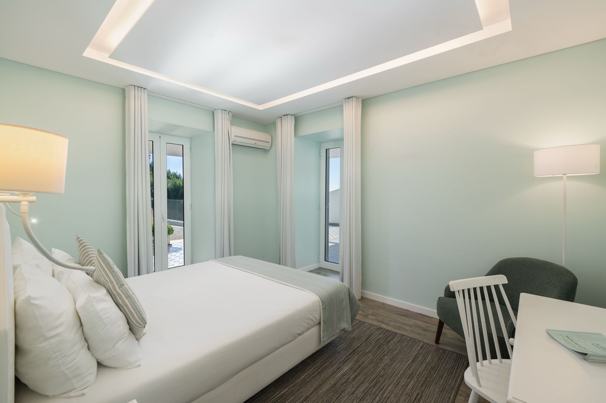 A hotel room with a bed, a desk, a chair, and a balcony with curtains.
