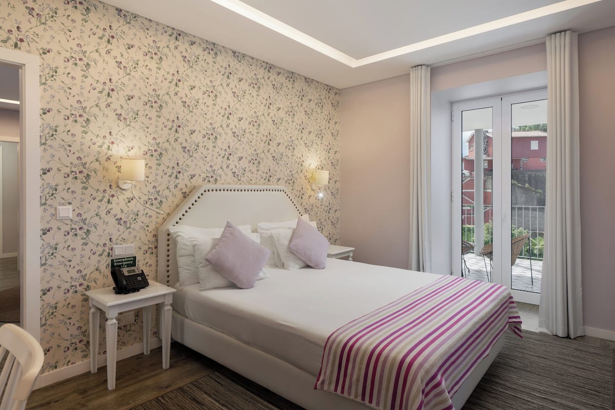 A hotel room with a floral wallpaper, a bed with white bedding and purple pillows, a bedside table with a phone, and a view of the outside through a sliding door.