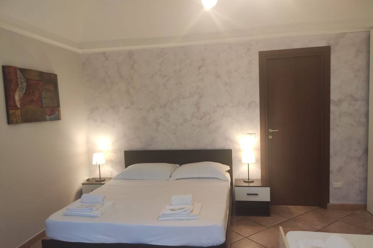 A neatly made bed with white linens and towels, two bedside lamps, a door, and a painting on the wall.