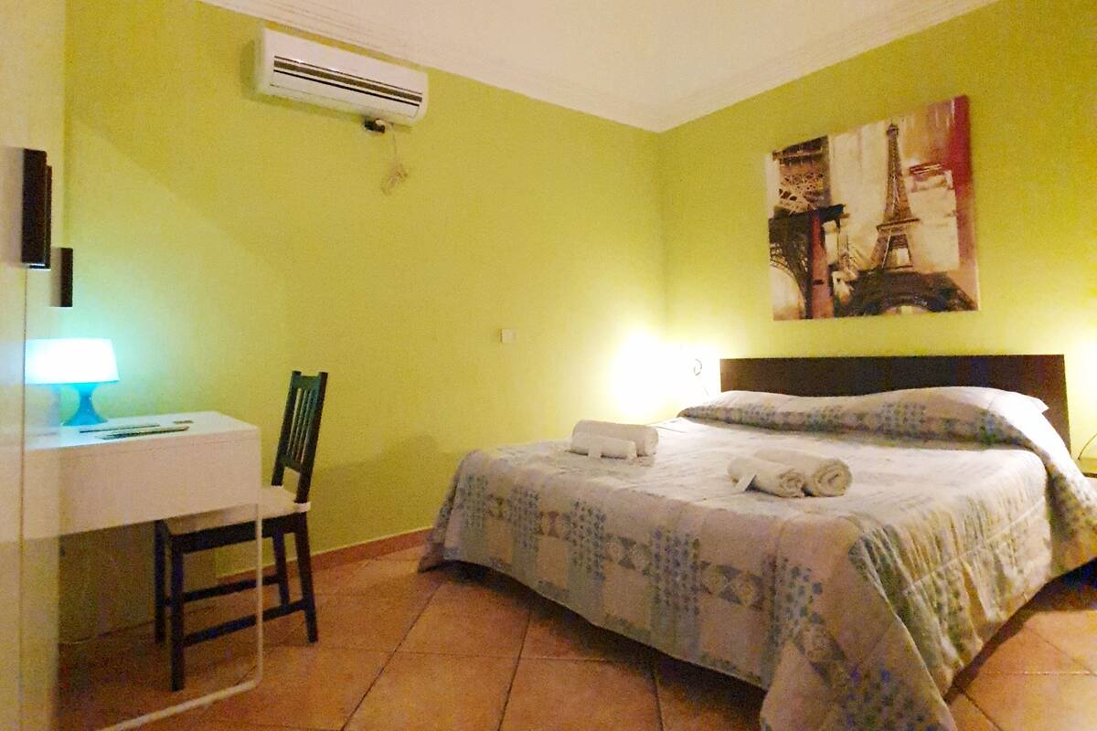 A bedroom with a bed, a desk with a chair, a lamp, and an air conditioner on the wall.