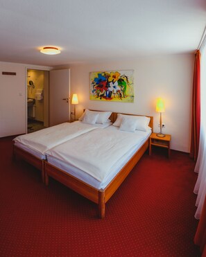 Une chambre d’hôtel avec un grand lit, une moquette rouge, deux lampes de chevet et un tableau abstrait aux couleurs vives accroché au mur.