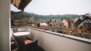 Un balcon avec une table et des chaises, donnant sur un quartier résidentiel avec des maisons et une colline boisée.