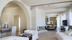 Una sala de estar moderna con sofás blancos, una mesa de centro y un televisor. Hay un comedor con una mesa y sillas. La habitación tiene un techo abovedado y suelo de madera.