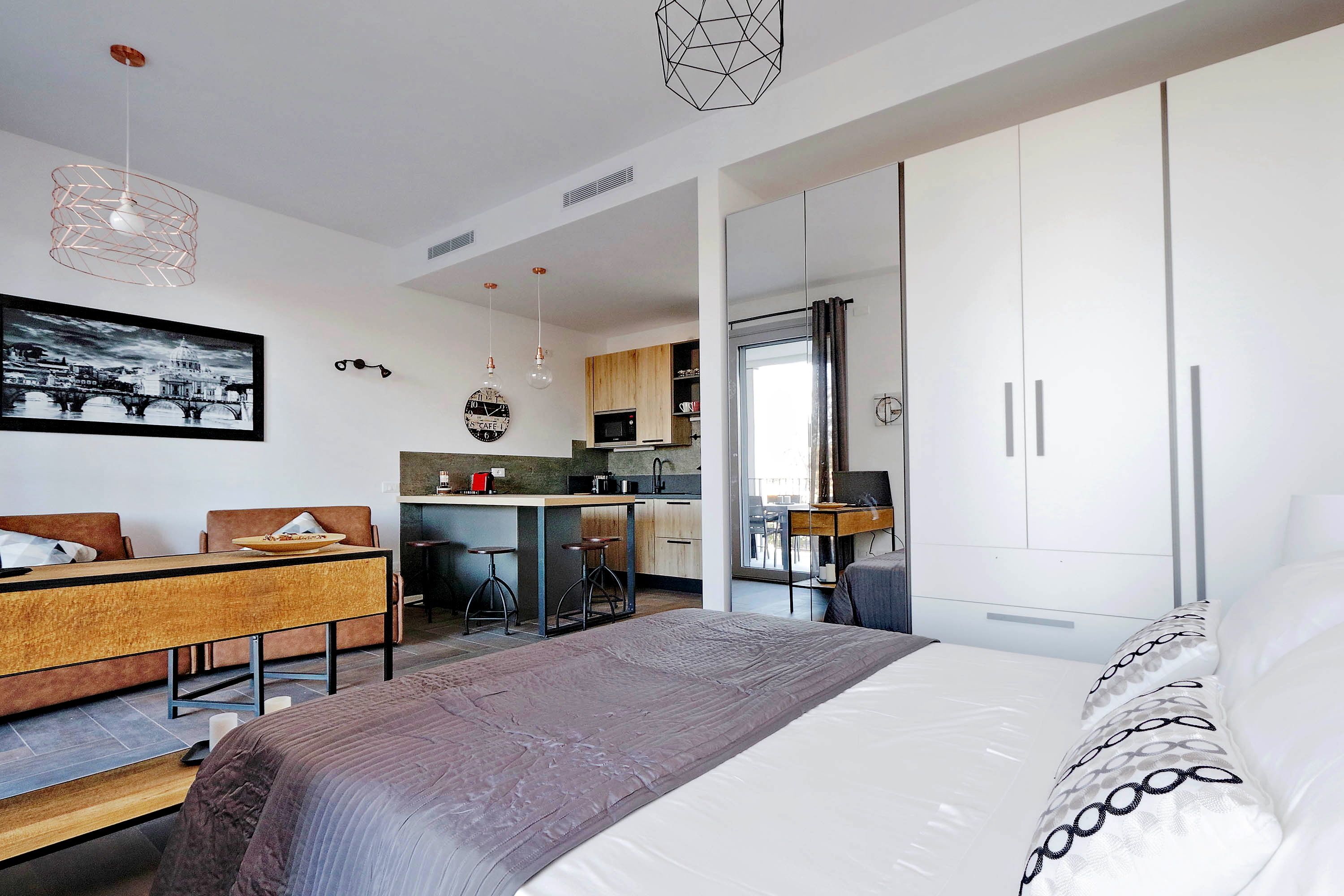 Une chambre d’hôtel moderne dotée d’un grand lit, d’un coin repas avec une table et des chaises en bois, d’une kitchenette et d’une grande armoire.