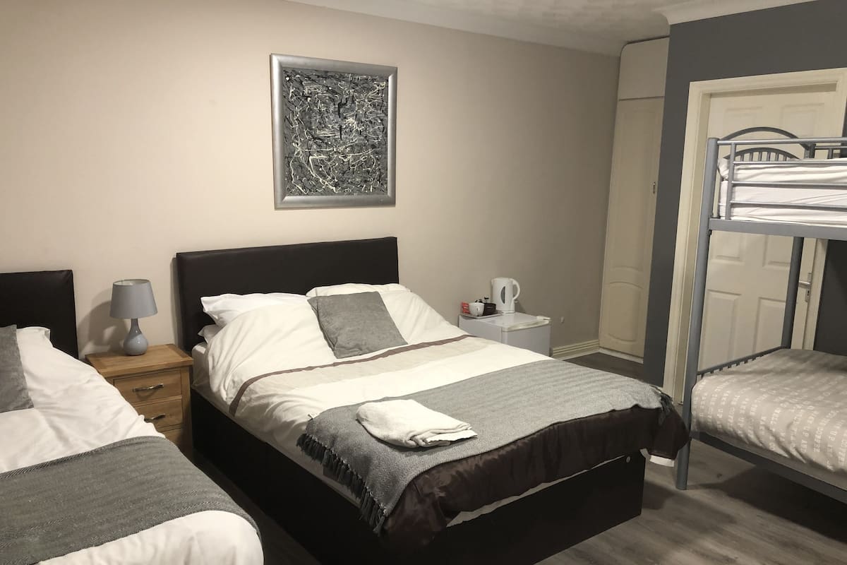 Quarto de hotel com duas camas, uma mesinha de cabeceira com abajur, uma obra de arte abstrata emoldurada na parede e uma cama beliche no canto.