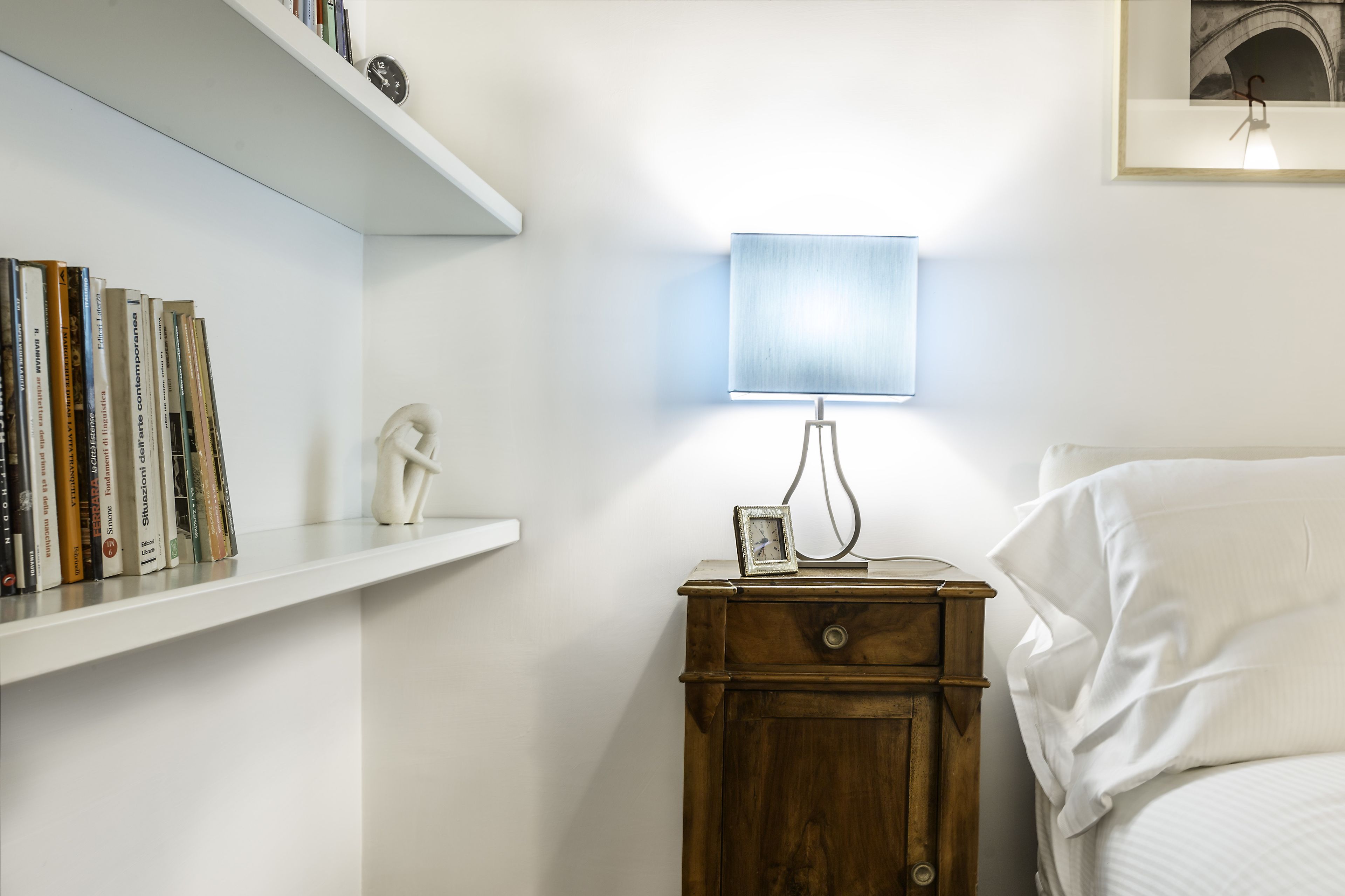 Un dormitorio con una mesita de noche, una lámpara, un reloj de noche, un cuadro en la pared y una estantería con libros.