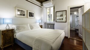 A bedroom with a large bed, a wooden nightstand, a bedside lamp, a radiator, a window, a mirror, and a door leading to a bathroom. - Luxury Flat in the Center of Rome (Rome)