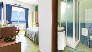 Ett hotellrum med en säng, ett skrivbord med en stol, ett litet bord med en växt, ett fönster med gardiner, ett badrum med dusch och ett handfat.