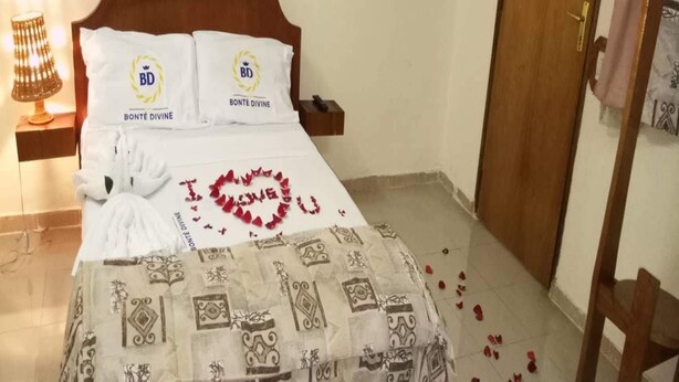 A neatly made bed with a heart-shaped decoration and scattered rose petals, a bedside lamp, and a wooden headboard with a power outlet. - Résidence Bonté Divine (Abidjan)