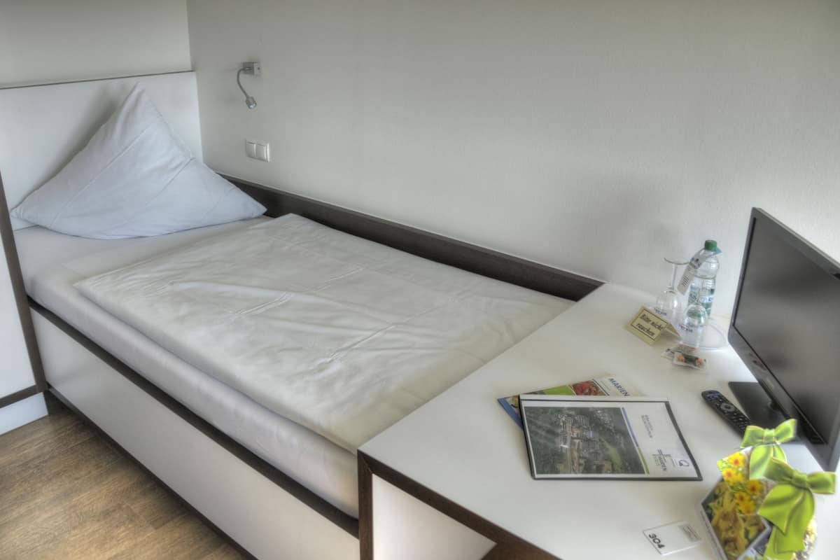 A single bed with a white mattress and pillow, a bedside table with a remote control, a water bottle, a magazine, and a small decorative item.