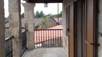 A stone balcony with a metal railing, a wooden door, and a view of a red-tiled roof in the background.