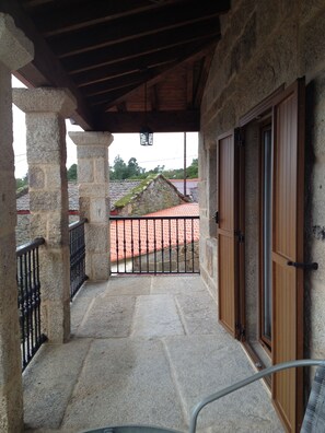 A stone balcony with a metal railing, a wooden door, and a view of a red-tiled roof in the background. - Alojamiento Rural Pazos (San Cristovo de Cea)