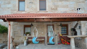 A stone building with a tiled roof, a wooden bench, two hanging chairs, a lawnmower, and a metal bucket. - Alojamiento Rural Pazos (San Cristovo de Cea)