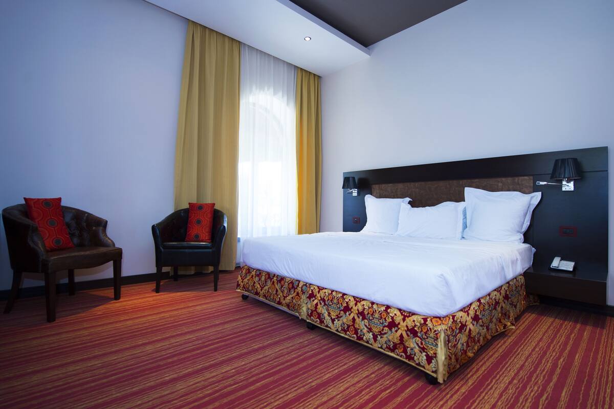 A hotel room with a large bed, two armchairs, a nightstand, and a window with curtains.