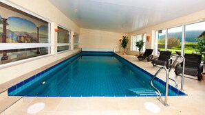 An indoor swimming pool with clear glass walls, a view of a mountainous landscape, and sun loungers along the side. - Hotel zum Postillion (Reit im Winkl)