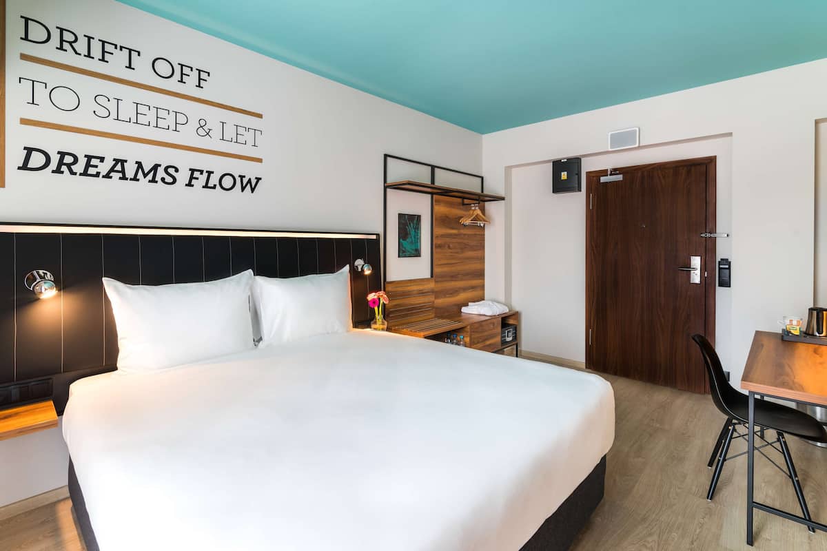 A hotel room with a large bed, a desk, a chair, and a wardrobe. There is a sign on the wall that says "DRIFT OFF TO SLEEP & LET DREAMS FLOW".