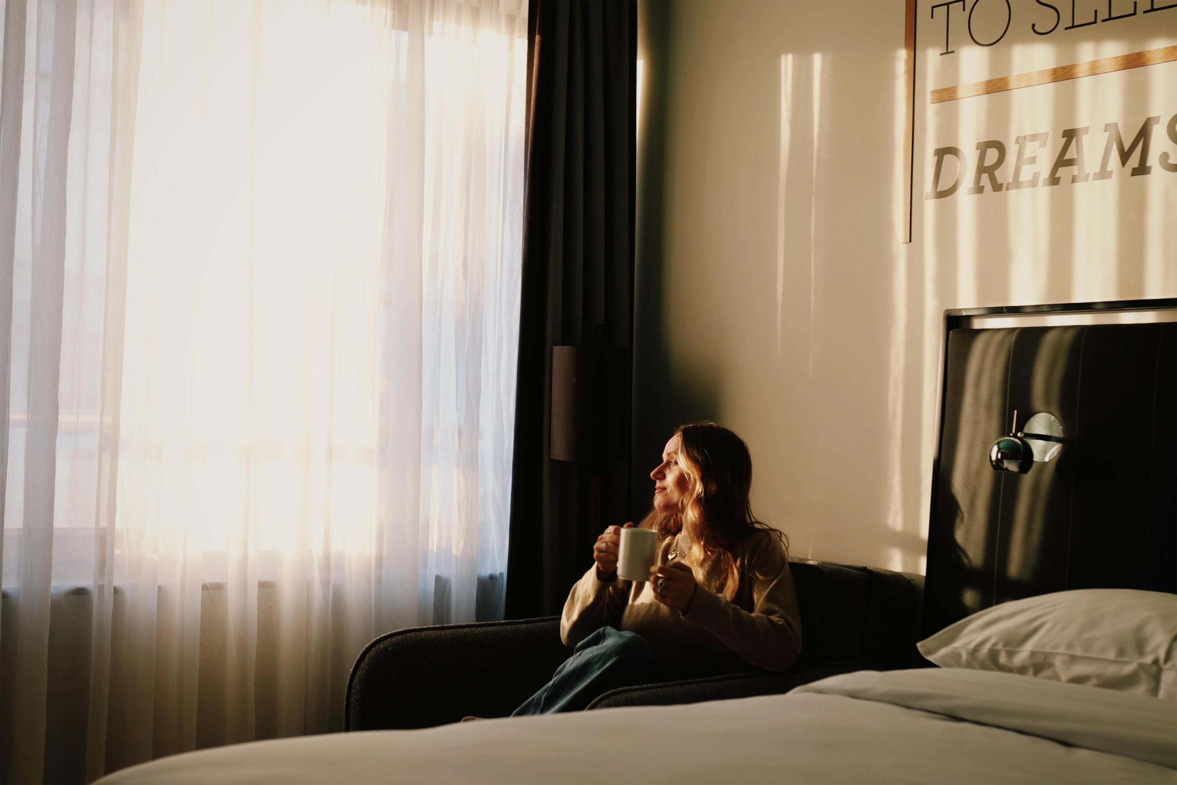 A person sitting on a bed in a hotel room with a window, a headboard, and a wall with text.